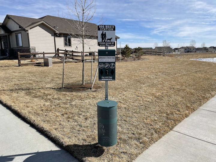 Dog waste station with disposal bin and bags on a grassy area near a sidewalk and houses.