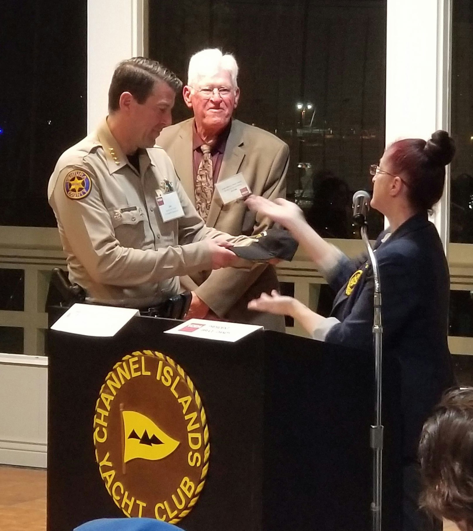 A speaker presents a hat to a male officer at an event with a backdrop featuring the Channel Islands Yacht Club logo.