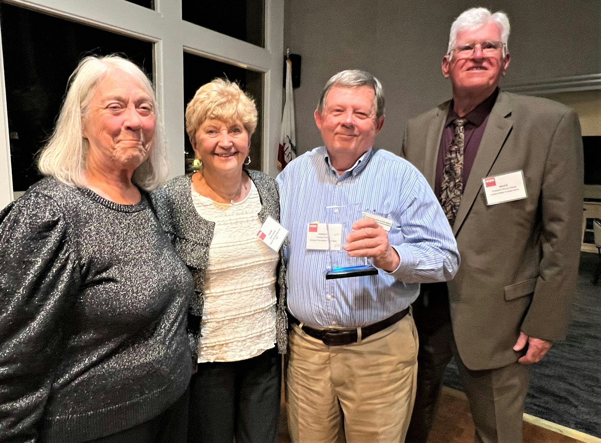 A group of four people smiling together at an event, one holding an award or plaque.