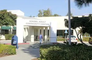 The image shows the entrance of the Goebel Adult Community Center, with a person walking in and a mailbox nearby.