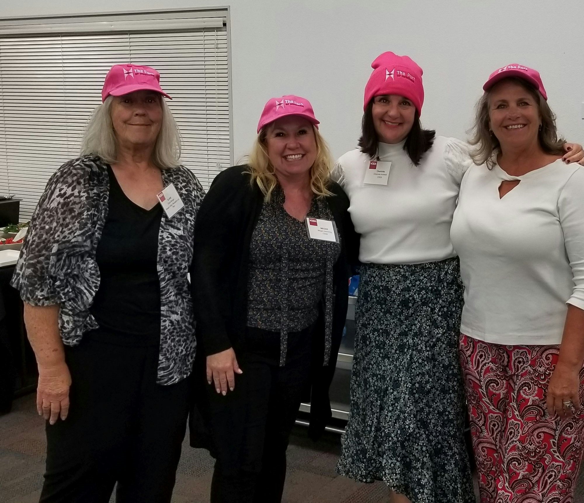 Four women, wearing pink hats, smile for the camera in a room with a casual setting. They seem to be part of an event or gathering.
