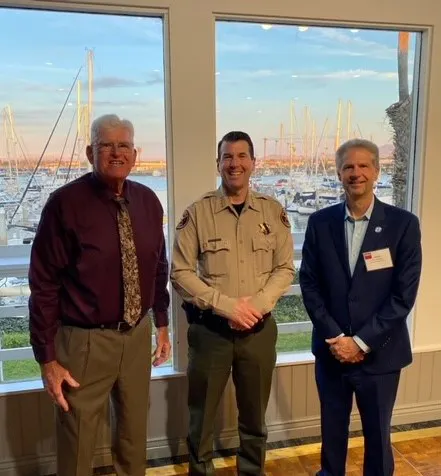 Three men standing indoors in front of large windows overlooking a marina with sailboats. The man on the left is wearing a maroon shirt and patterned tie, the man in the middle is in a tan uniform, and the man on the right is in a navy blue suit with a name badge.