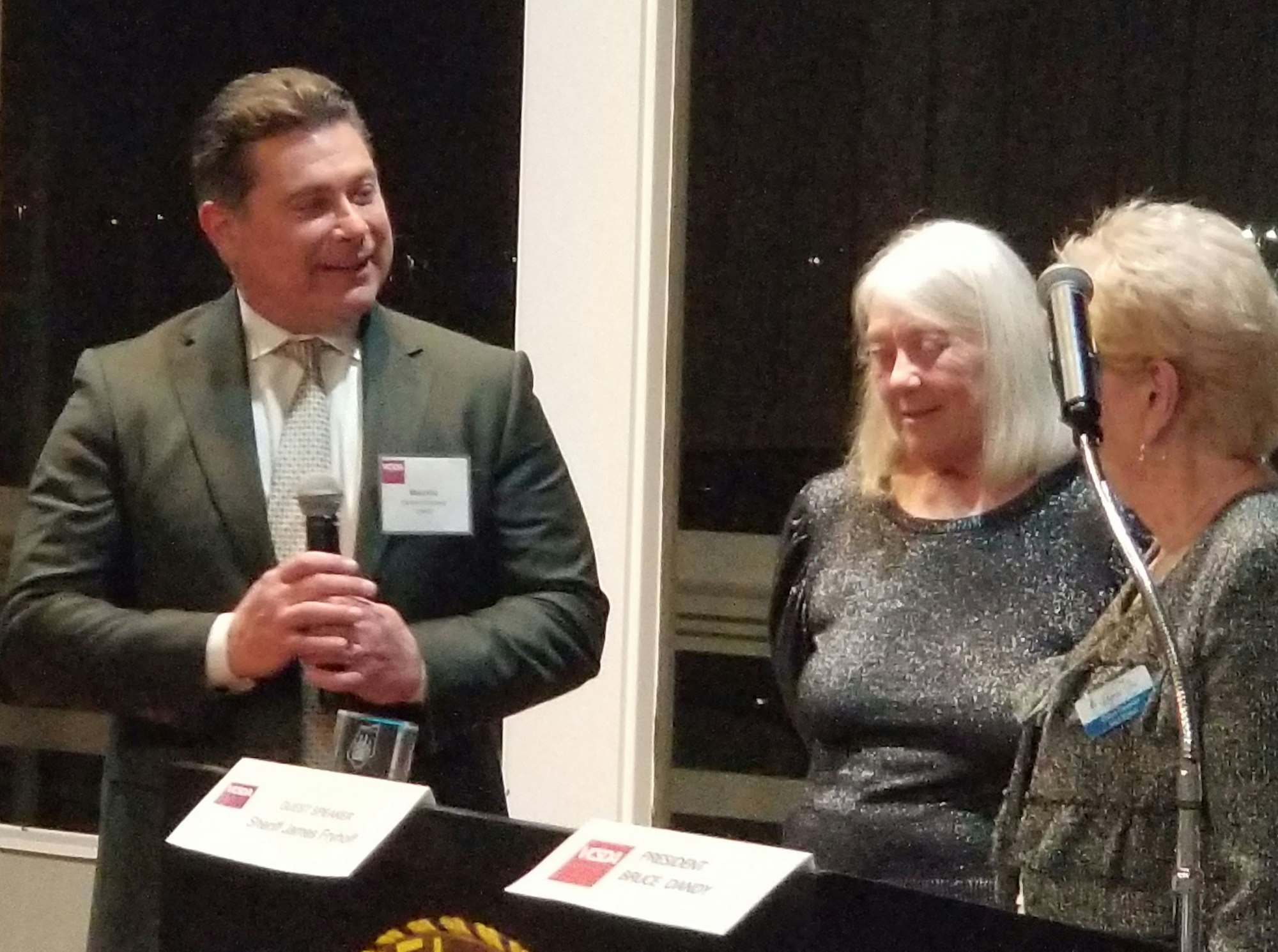 An event or gathering featuring a speaker at a podium with two women listening attentively, all dressed in formal attire.