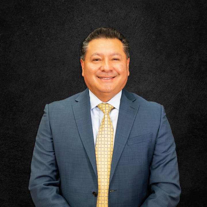 A person in a blue suit and yellow tie smiling against a dark background.