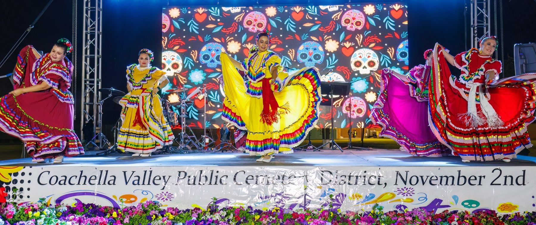 Dancers in colorful traditional dresses perform on stage with vibrant Day of the Dead-themed backdrop.