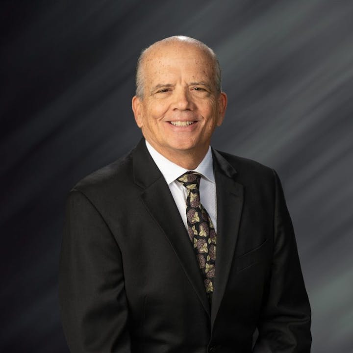 A man in a suit and tie smiles against a blurred dark background.