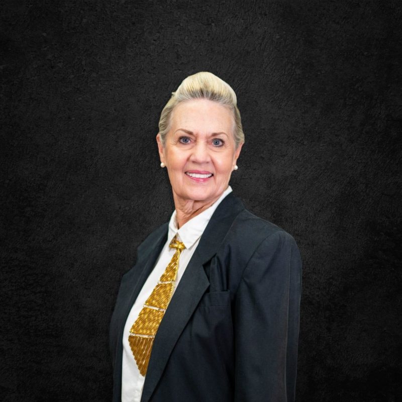 An older woman in a suit and patterned tie stands against a dark textured background.