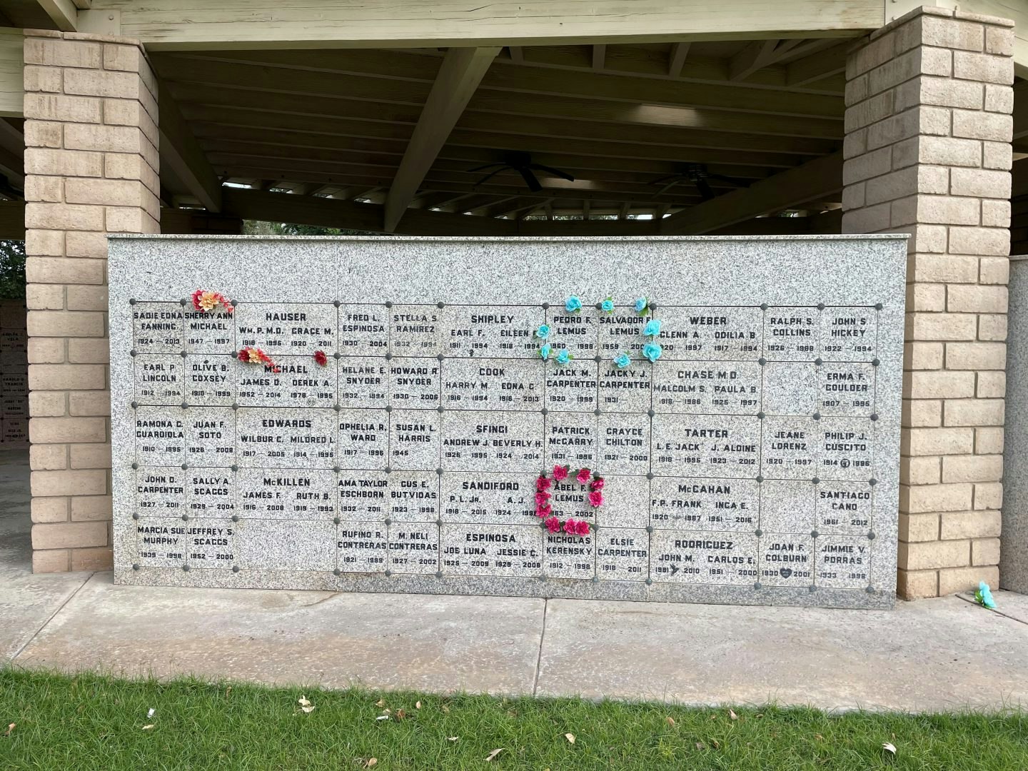 A granite wall with engraved names and dates, adorned with colorful artificial flowers, likely part of a memorial or columbarium.