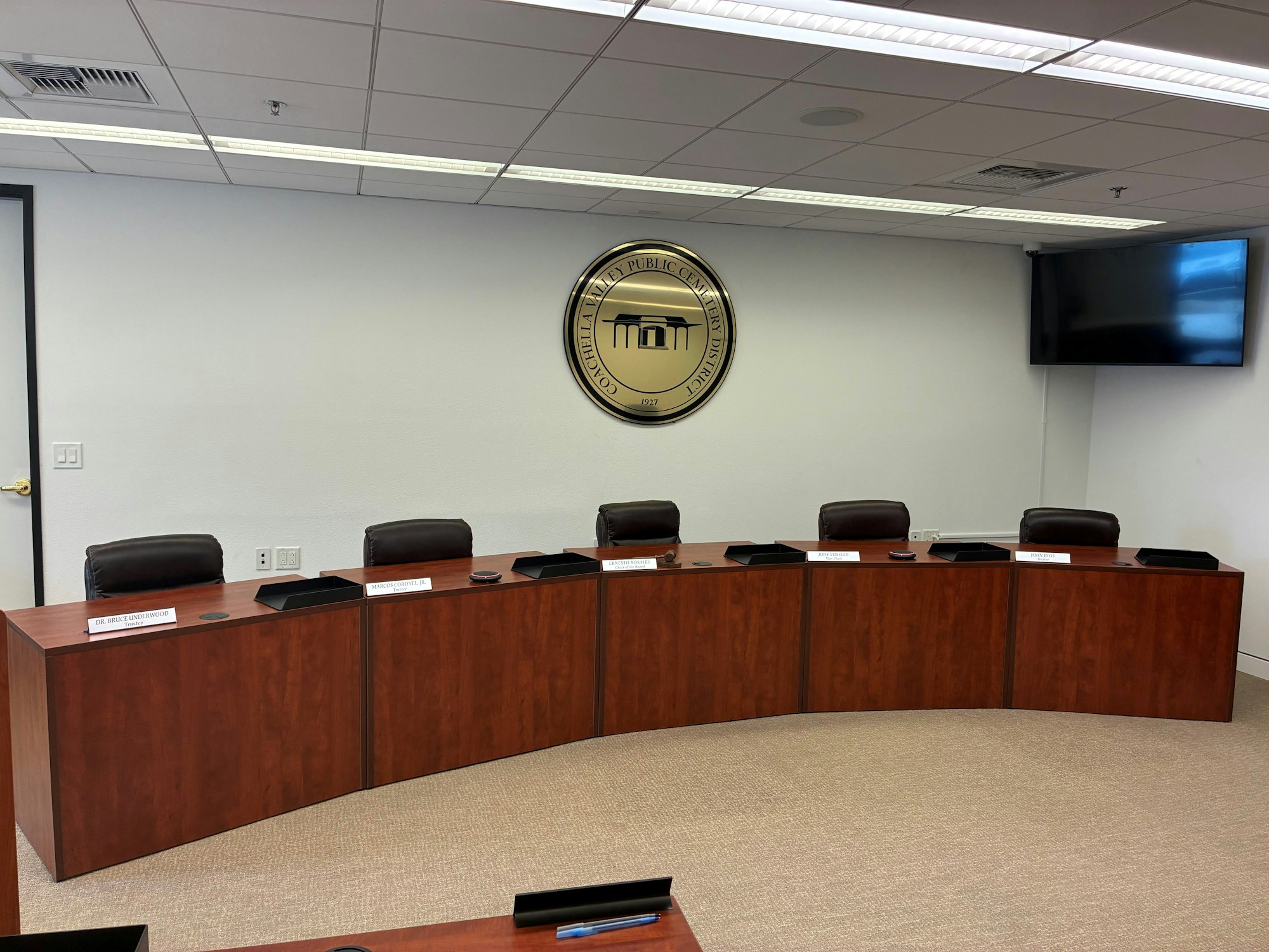 Meeting room with a curved wooden table, chairs, nameplates, a seal on the wall, and a TV screen.