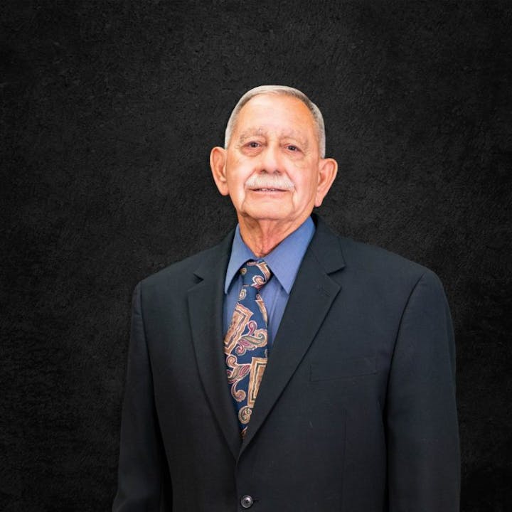 An elderly man in a suit and tie stands against a dark background.