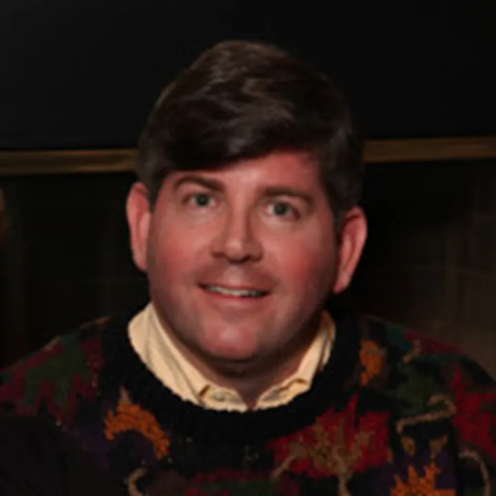 The image shows a man with short hair, wearing a colorful sweater, smiling in a cozy indoor setting.