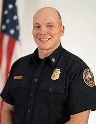 A person in uniform smiling, with a flag in the background.