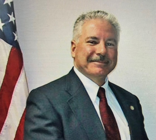 A man in a suit and tie smiling, with a U.S. flag in the background.