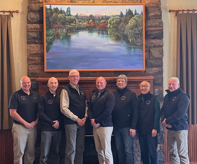 Seven men standing indoors, smiling, in front of a landscape painting of a lake and trees.