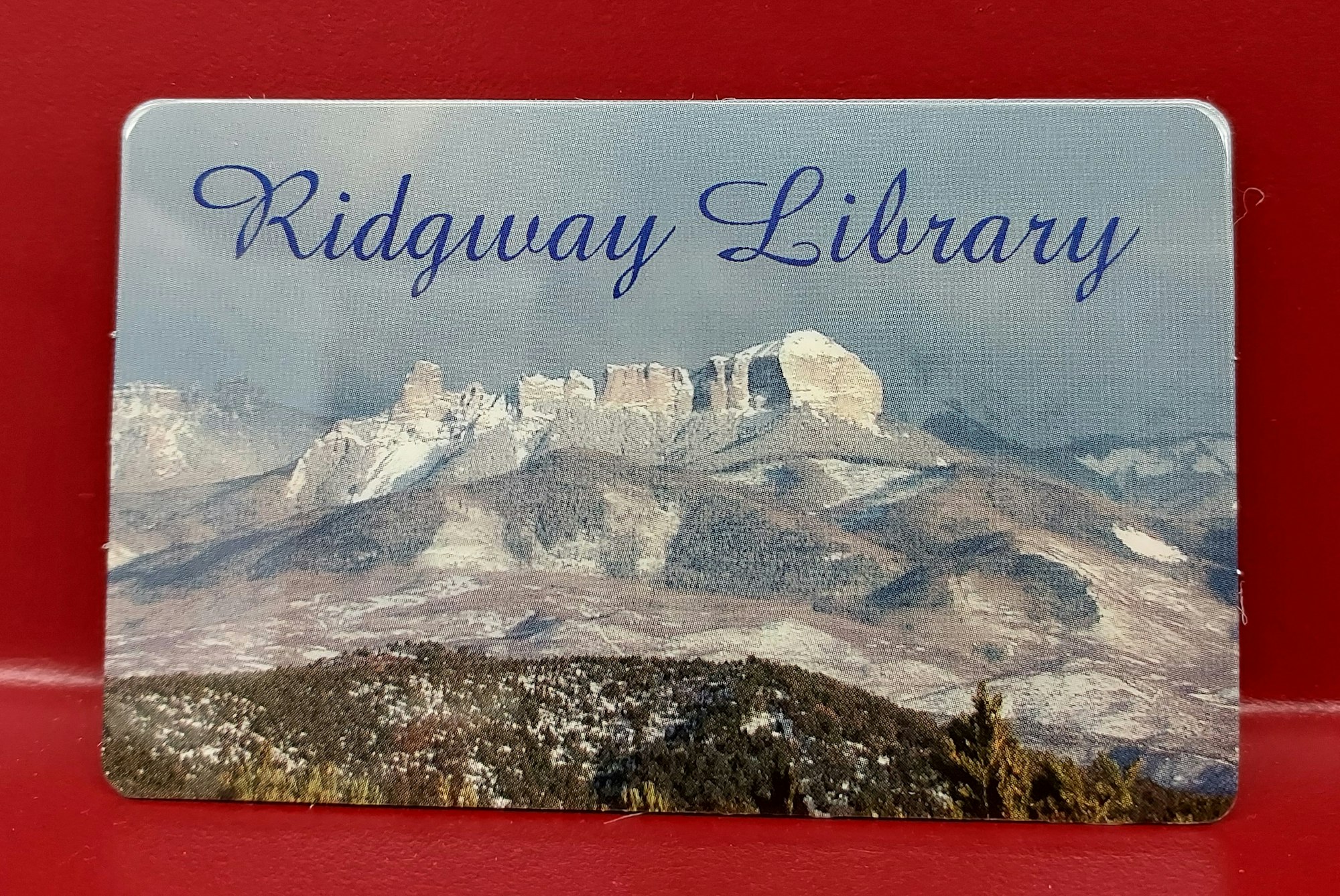 Ridgway Library card against a red background. Image on card is of Cimarron mountain range to east of Ridgway.