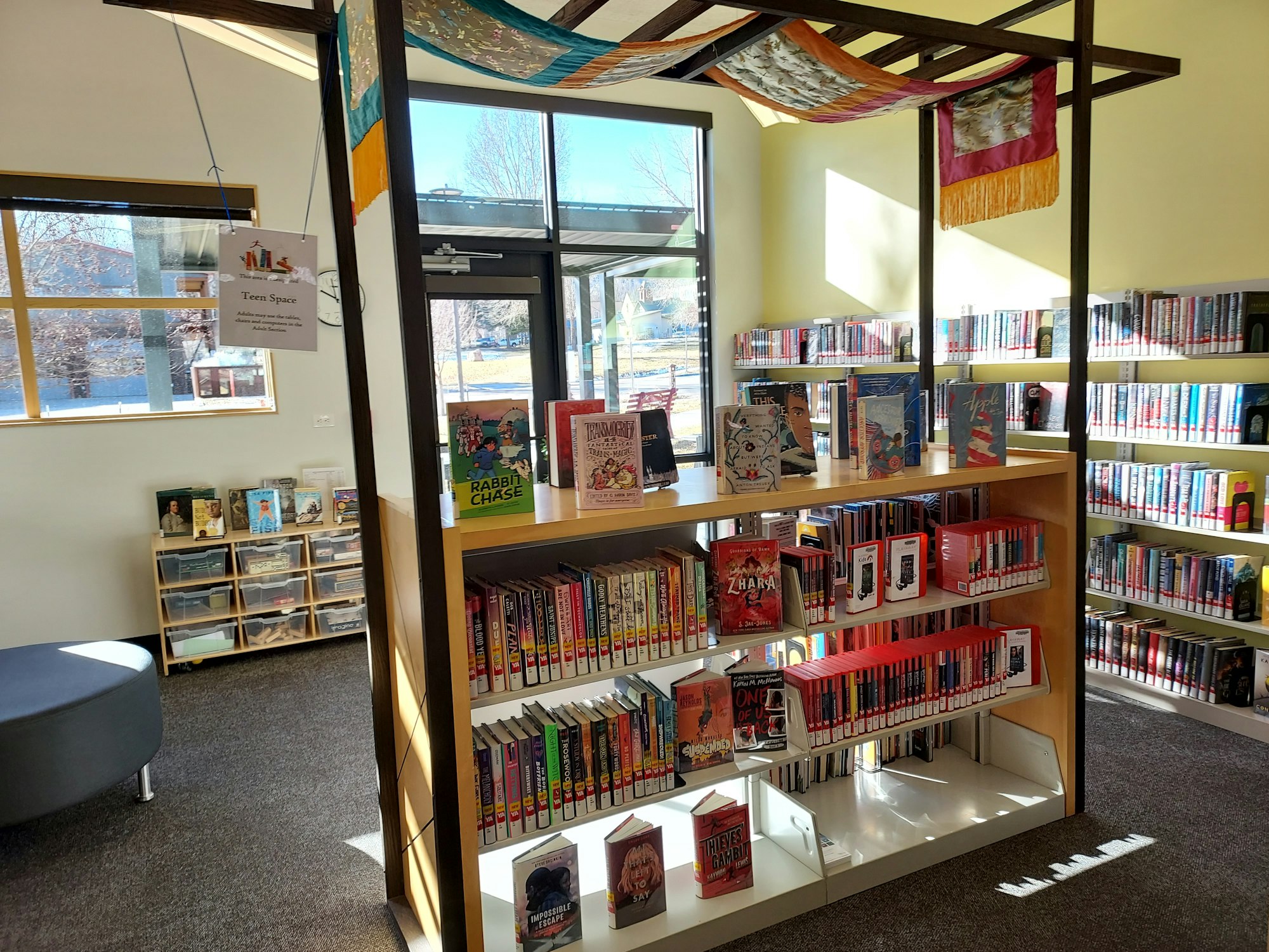 Photo of library teen area centered on new shelf with books and playaways on the shelves. More books are shelved along the wall. In back is a set of shelves and drawers with books on top and activities in the drawer. On the side is a round  ottoman for sitting.