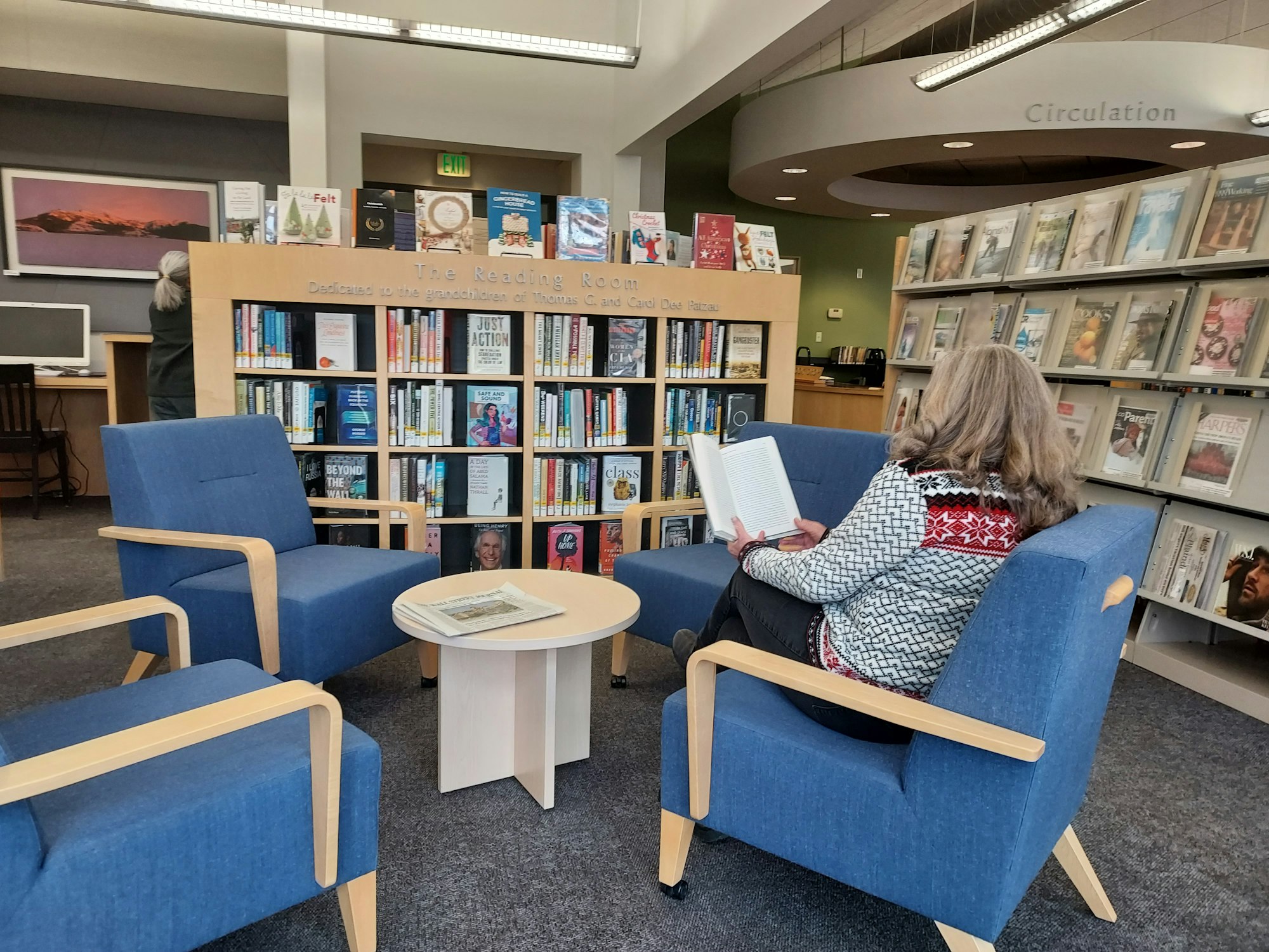 Reading area in the library. There are shelves with books and magazines. Two people are in the area, one sitting on a chair.