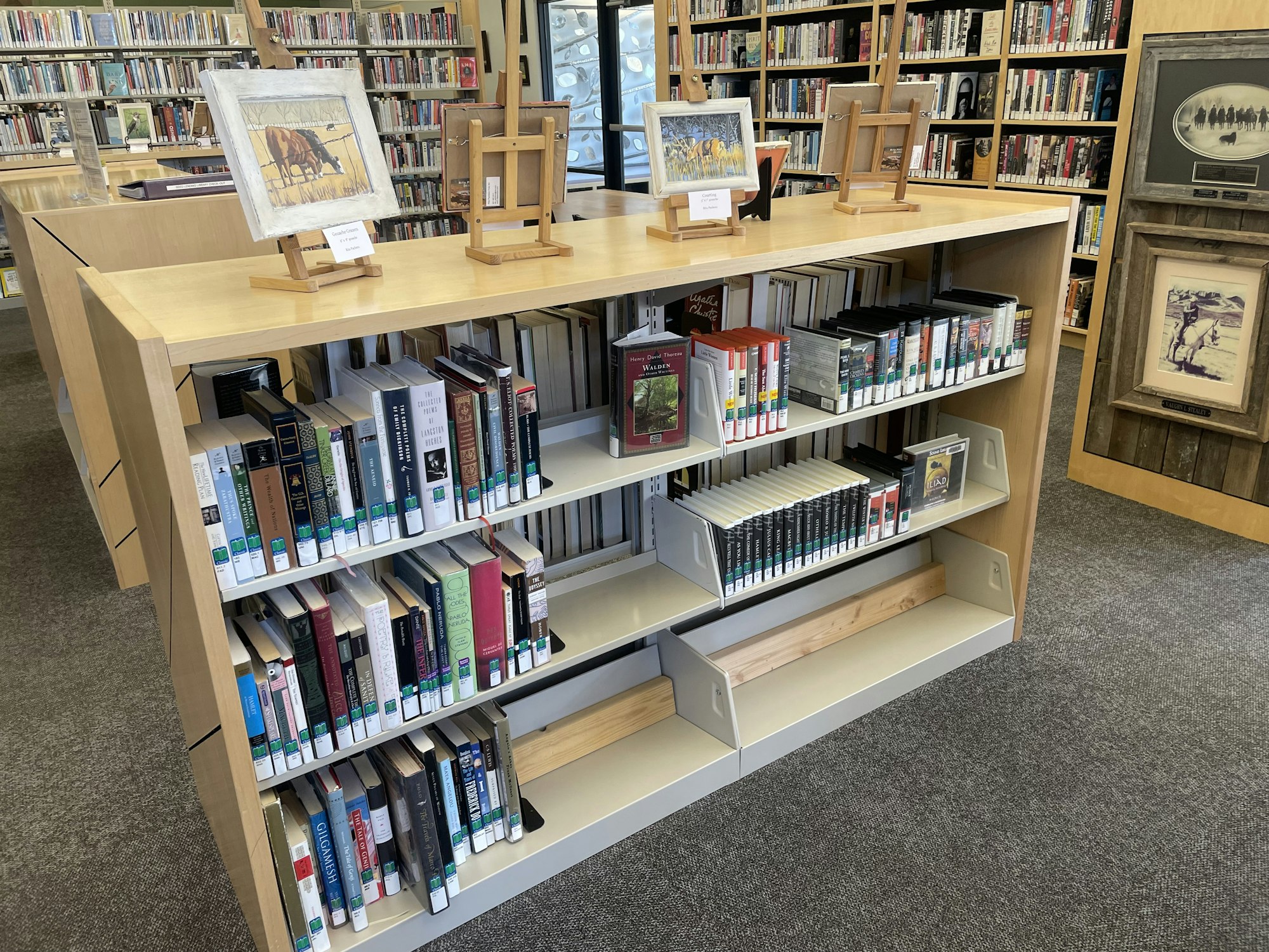 Photo of one side of Classics shelf at library, non fiction books, playaways, and books on CD on shelf. Artwork atop shelf and in background.