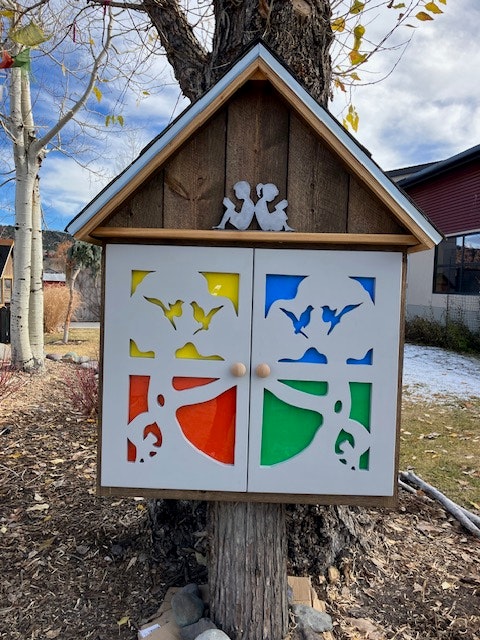 A colorful decorative little free library shaped like a house, featuring cut-out designs and vibrant panels in blue, green, yellow, and red.