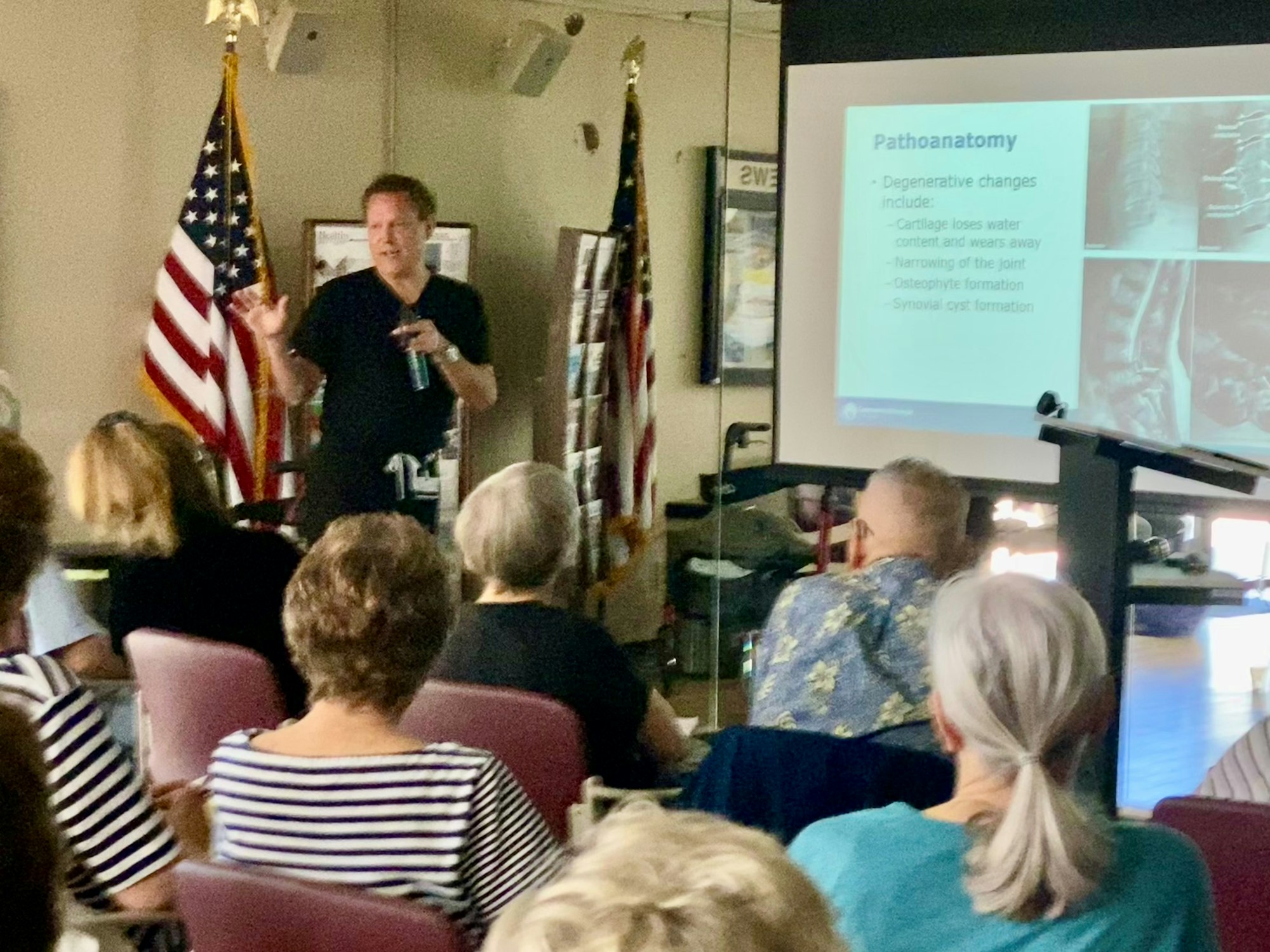 A presentation with an audience, a speaker discussing "Pathoanatomy", and U.S. flag visible in the background.