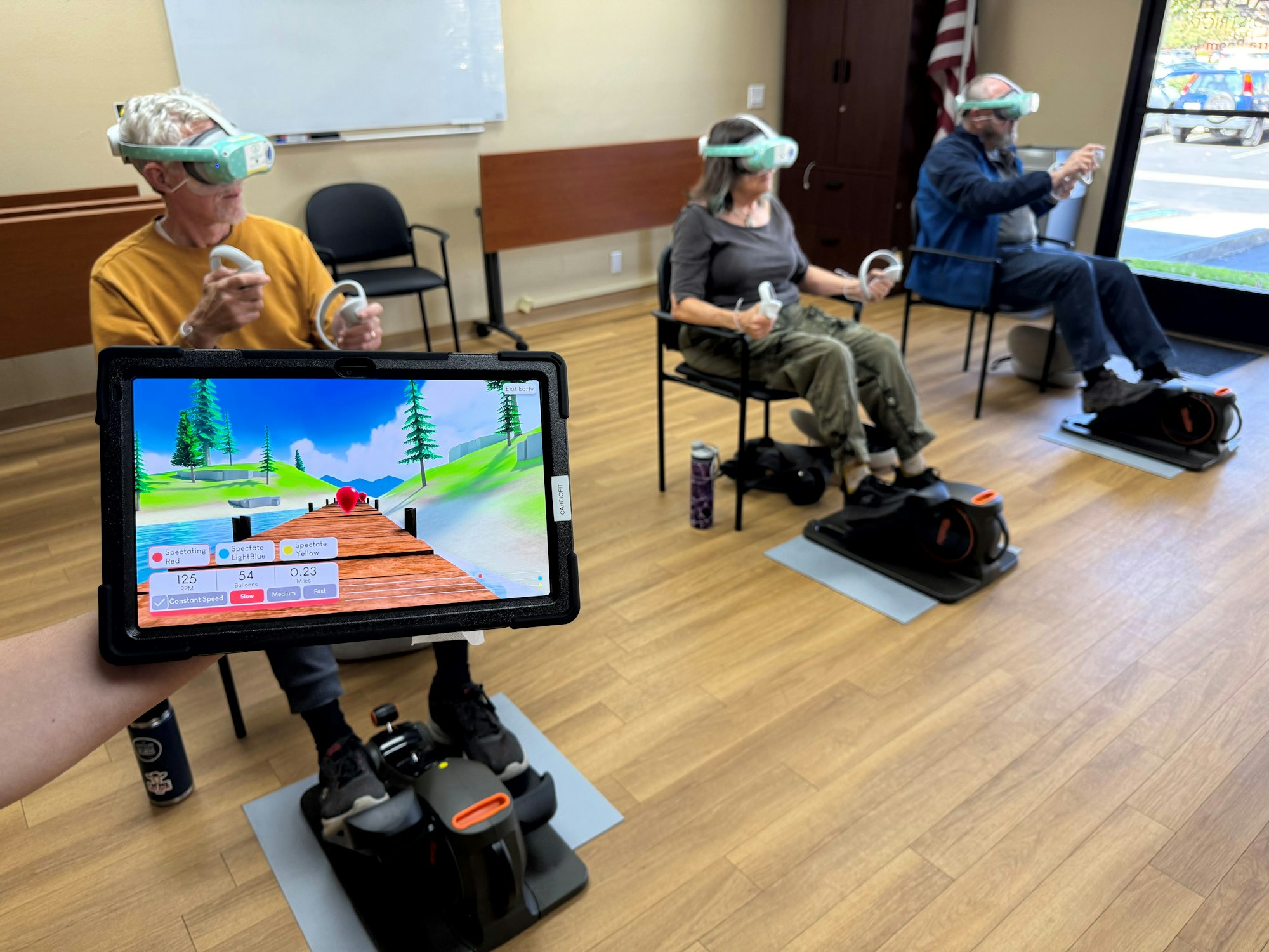 Three people using VR headsets and hand controllers while sitting on exercise machines, with a tablet displaying a skiing game.