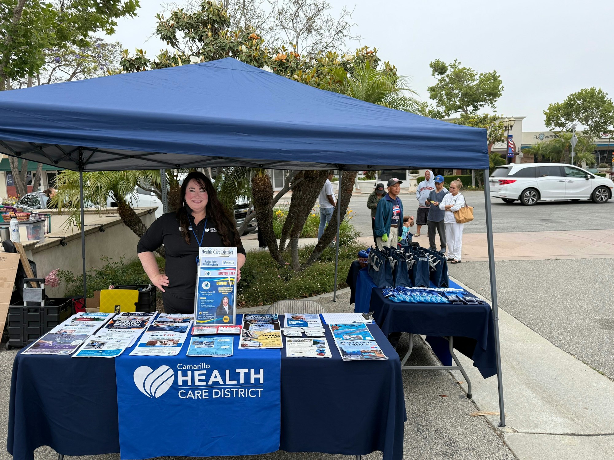 A woman stands in a blue tent with "Camarillo Health Care District" materials on tables, surrounded by trees and people in the background.
