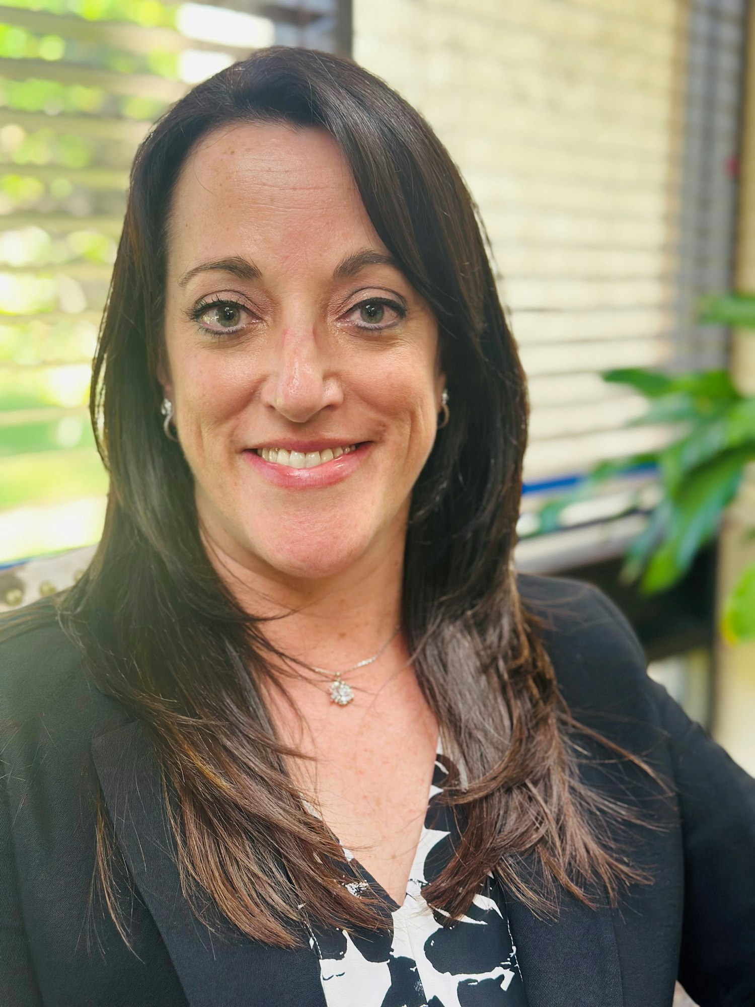 A person smiling, wearing a black blazer and patterned shirt, with a green, blurred background.