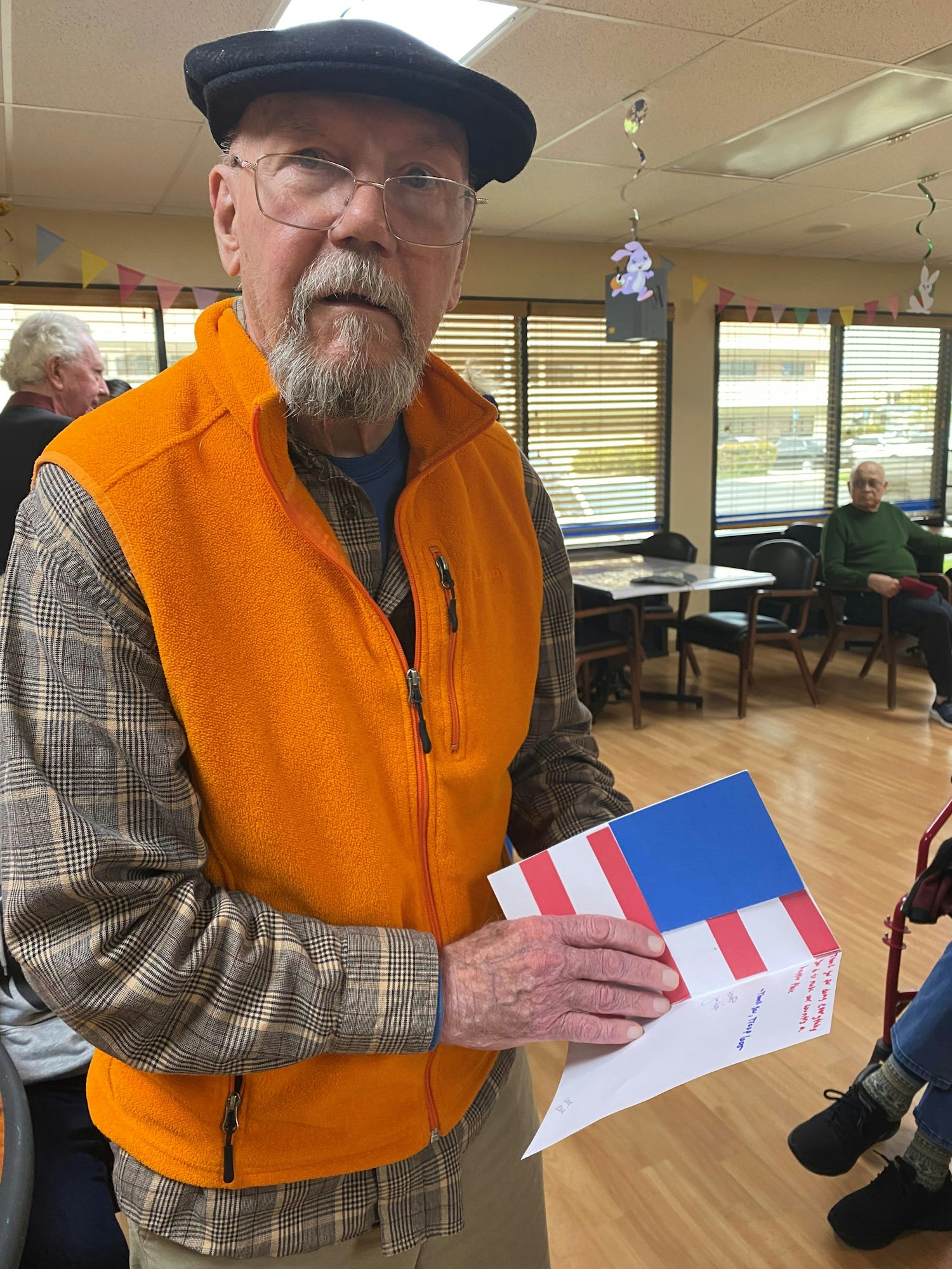 An elderly man holds a handmade card with red and white stripes and a blue section, standing in a decorated room.