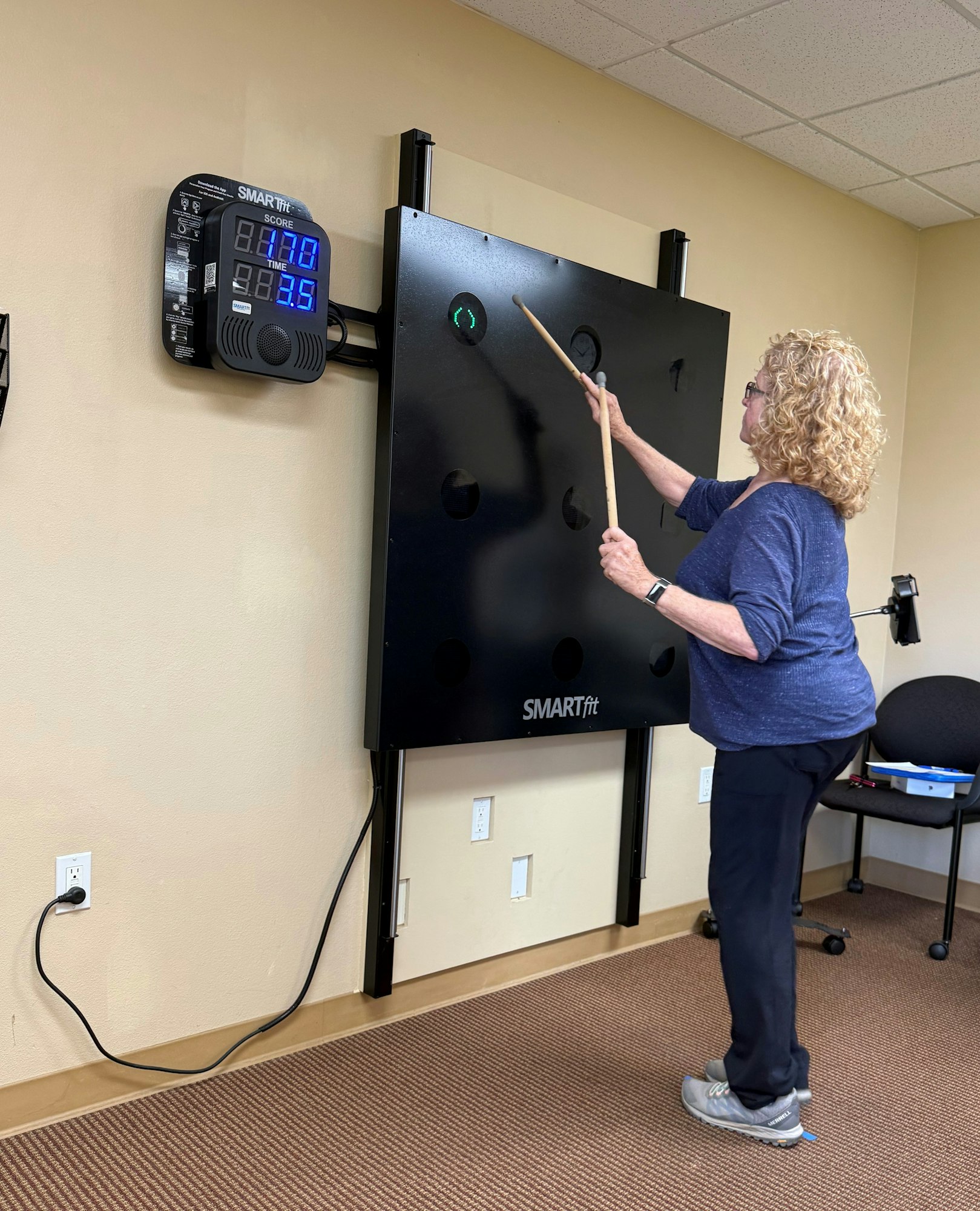An older woman is playing a SMARTfit game, hitting targets on a panel with a stick, focused on her score and time.