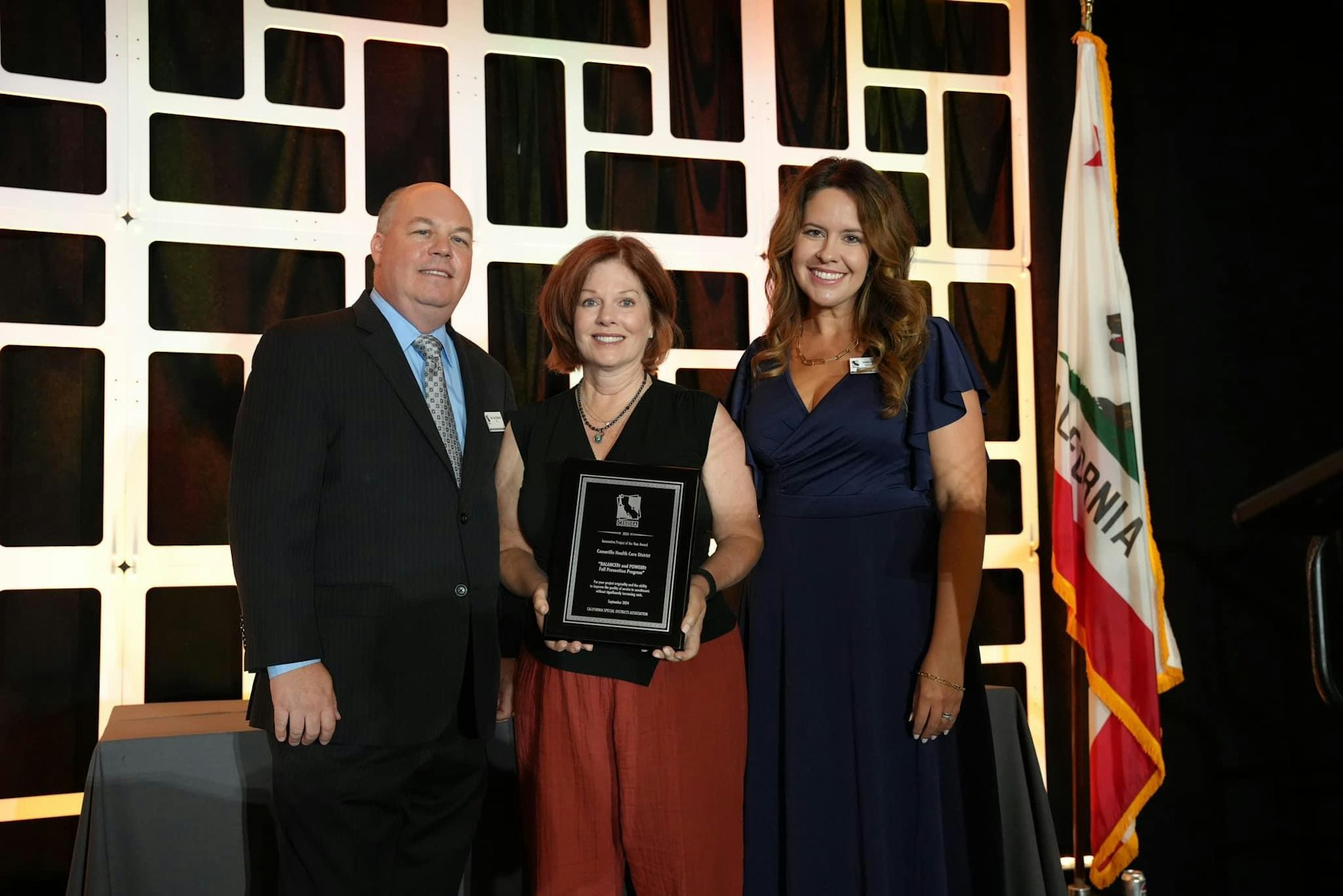 Three people posing with an award plaque at a formal event with a California flag in the background.