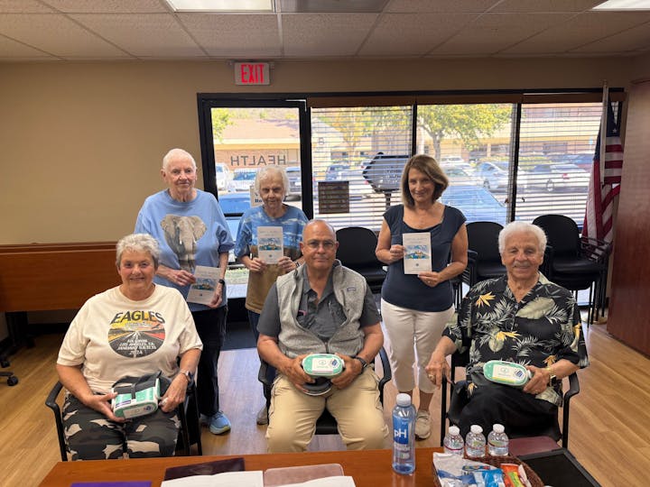 A group of six people posing indoors, holding pamphlets and items. Bottled water and snacks on the table in front of them.