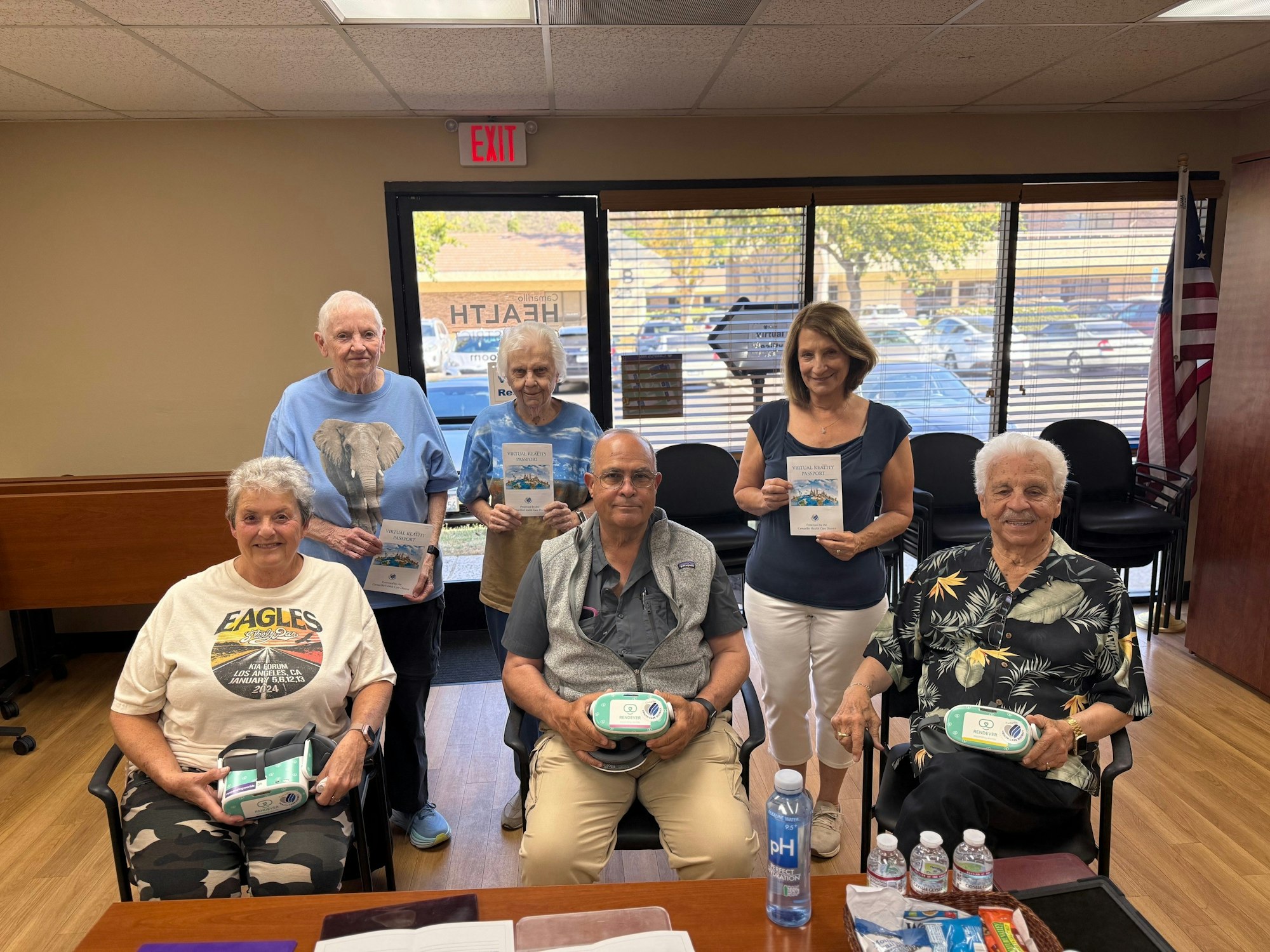 A group of six people posing indoors, holding pamphlets and items. Bottled water and snacks on the table in front of them.