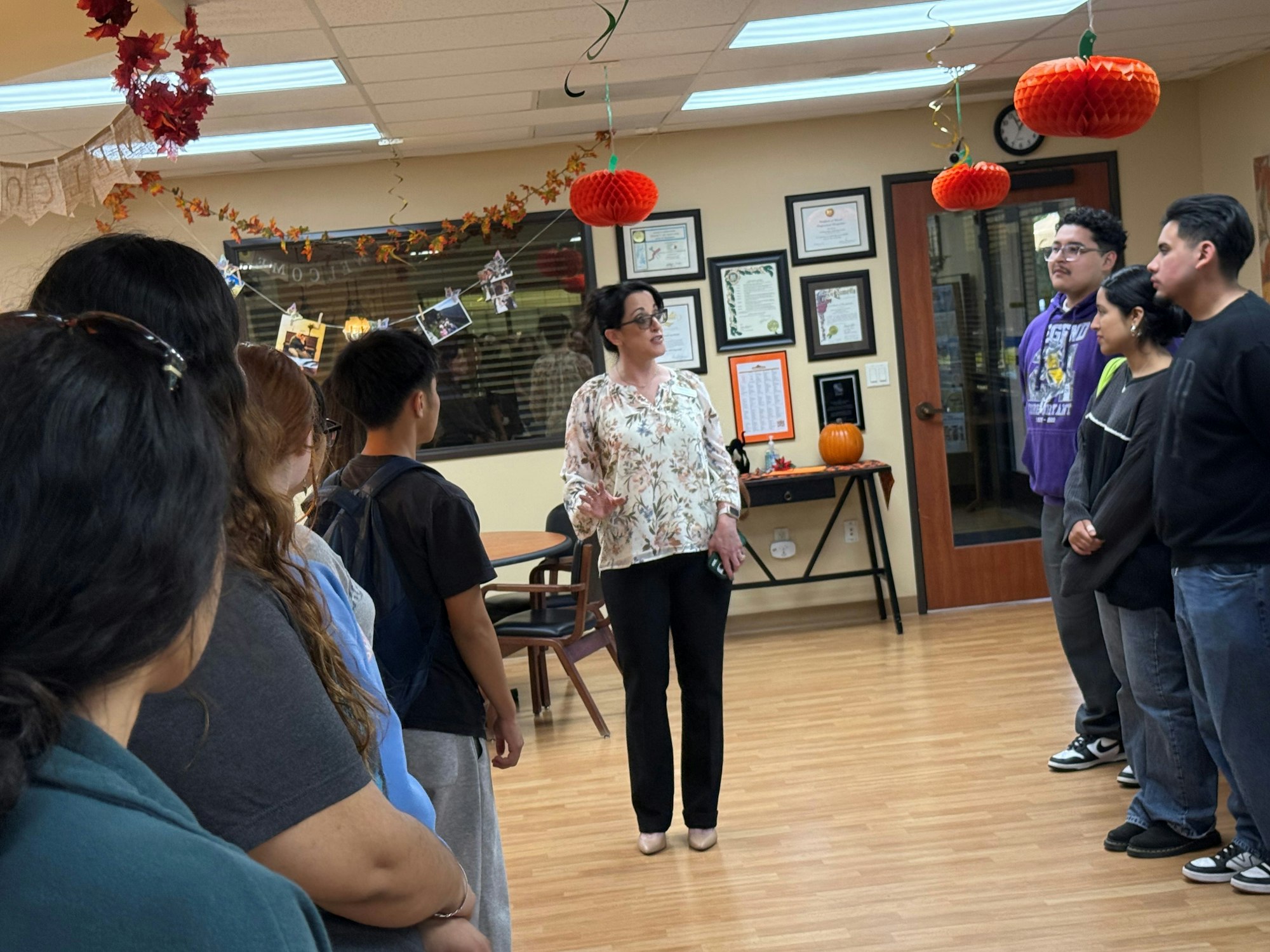 A group of people in a decorated room with hanging pumpkins, listening to a woman speaking.