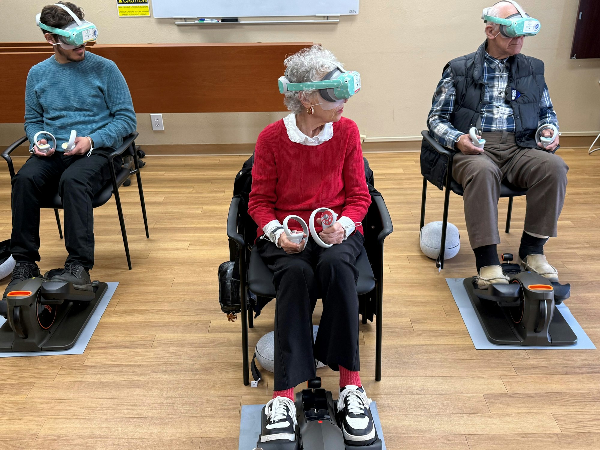 Three people seated wearing VR headsets, holding controllers, with foot pedals on the floor.