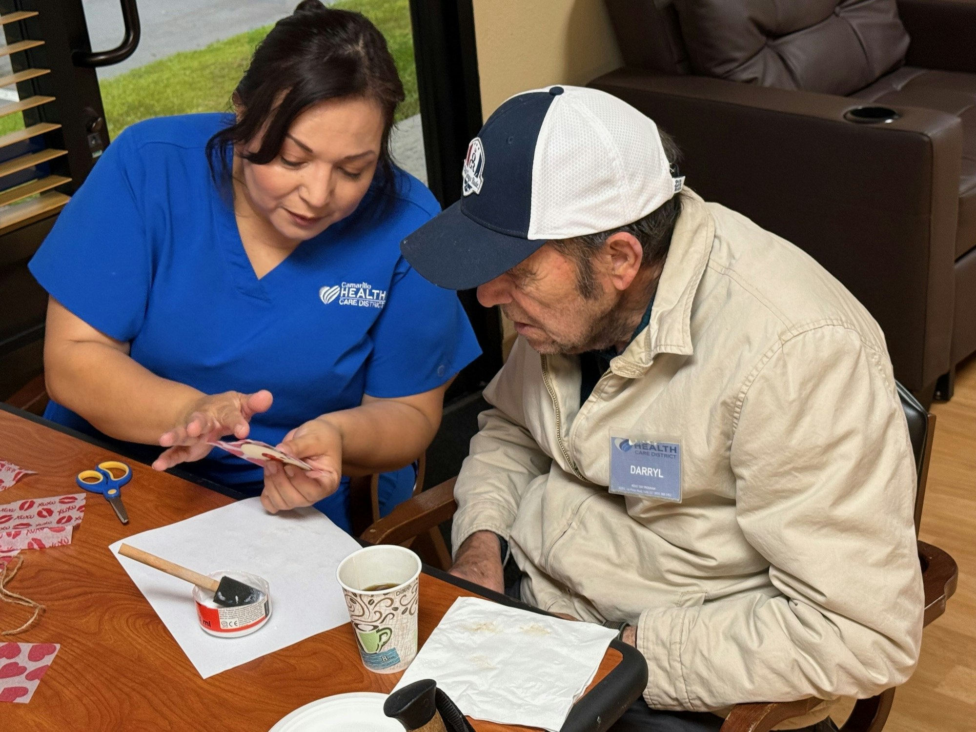 A healthcare worker shows something to an older man at a table with craft materials.