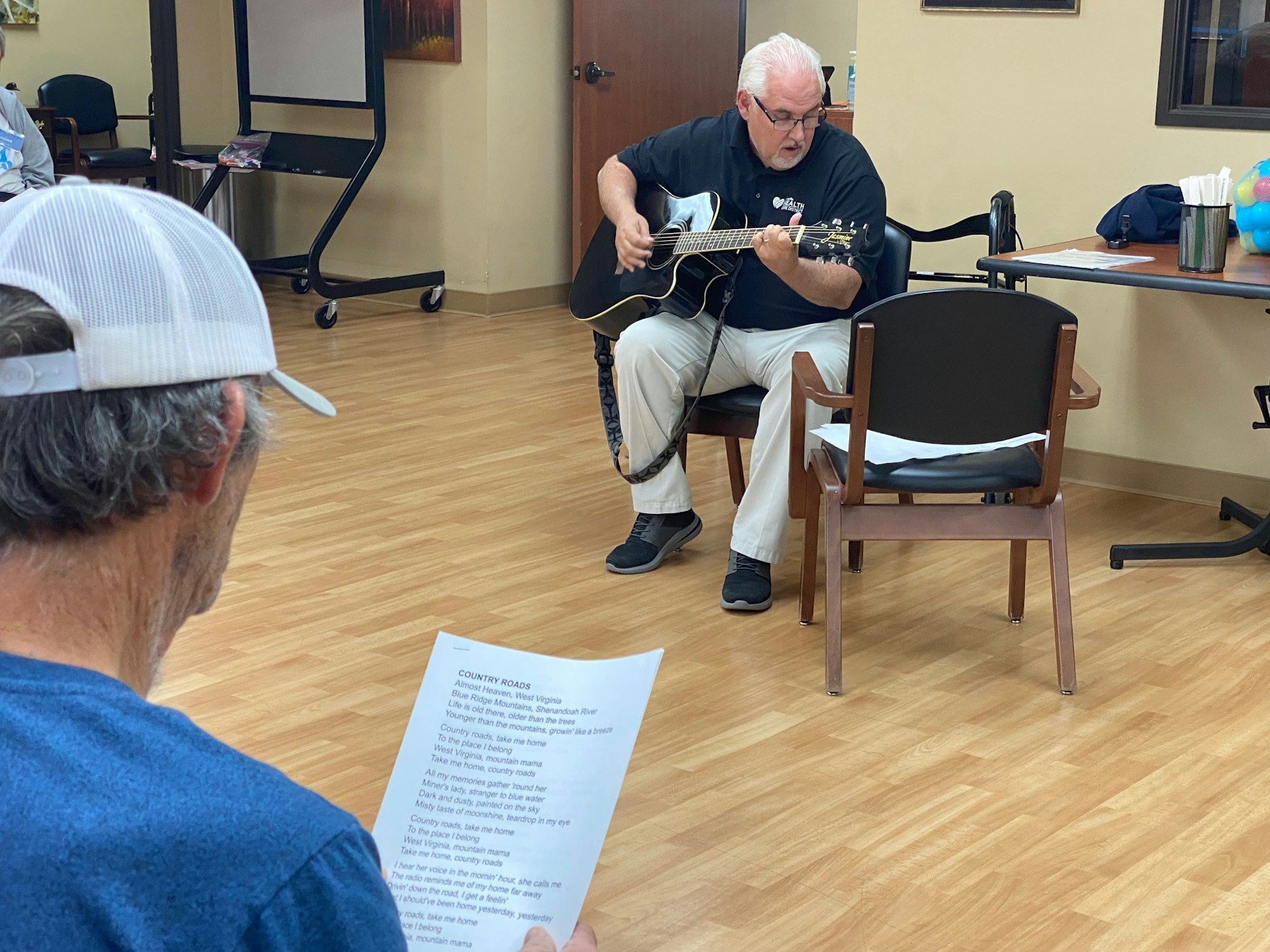 Man playing guitar for another with "Country Roads" lyrics sheet.