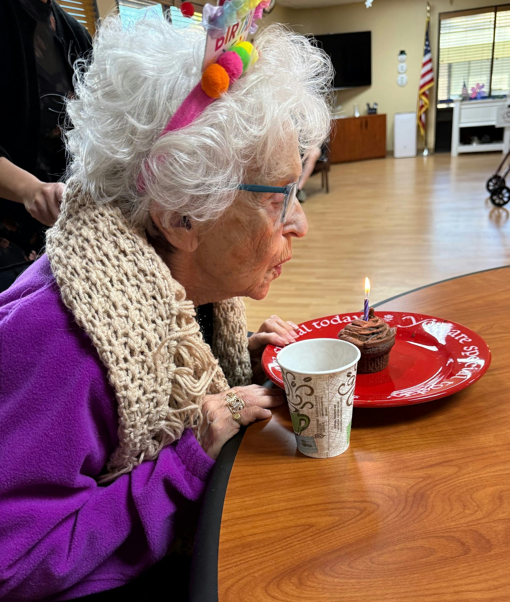An elderly woman with a "Birthday" headband blows out a candle on a cupcake. She holds a cup, celebrating at a table.