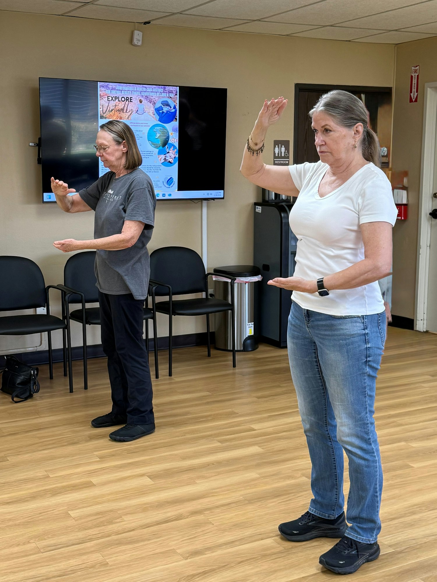Two women are practicing movements in a room with chairs and a TV displaying a poster.