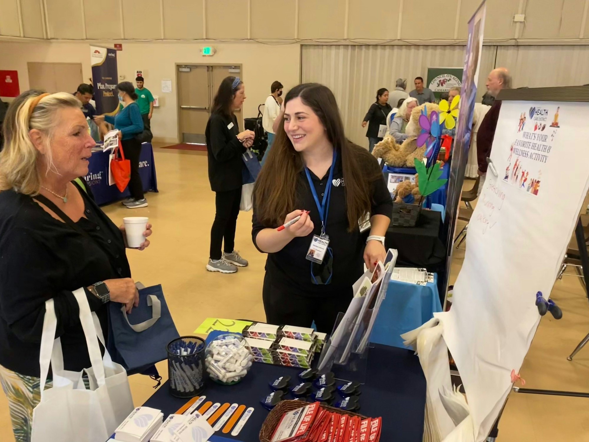 Two women interacting at an indoor booth with promotional materials and a wellness activity sign.