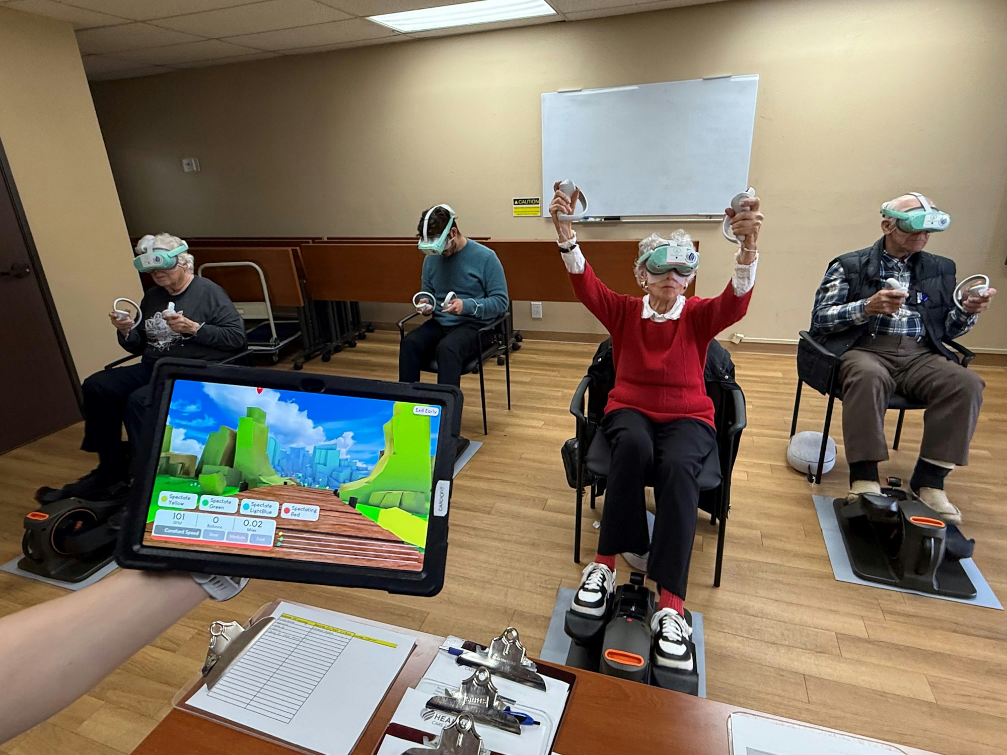 People wearing VR headsets using exercise equipment, viewed through a tablet showing a virtual environment.
