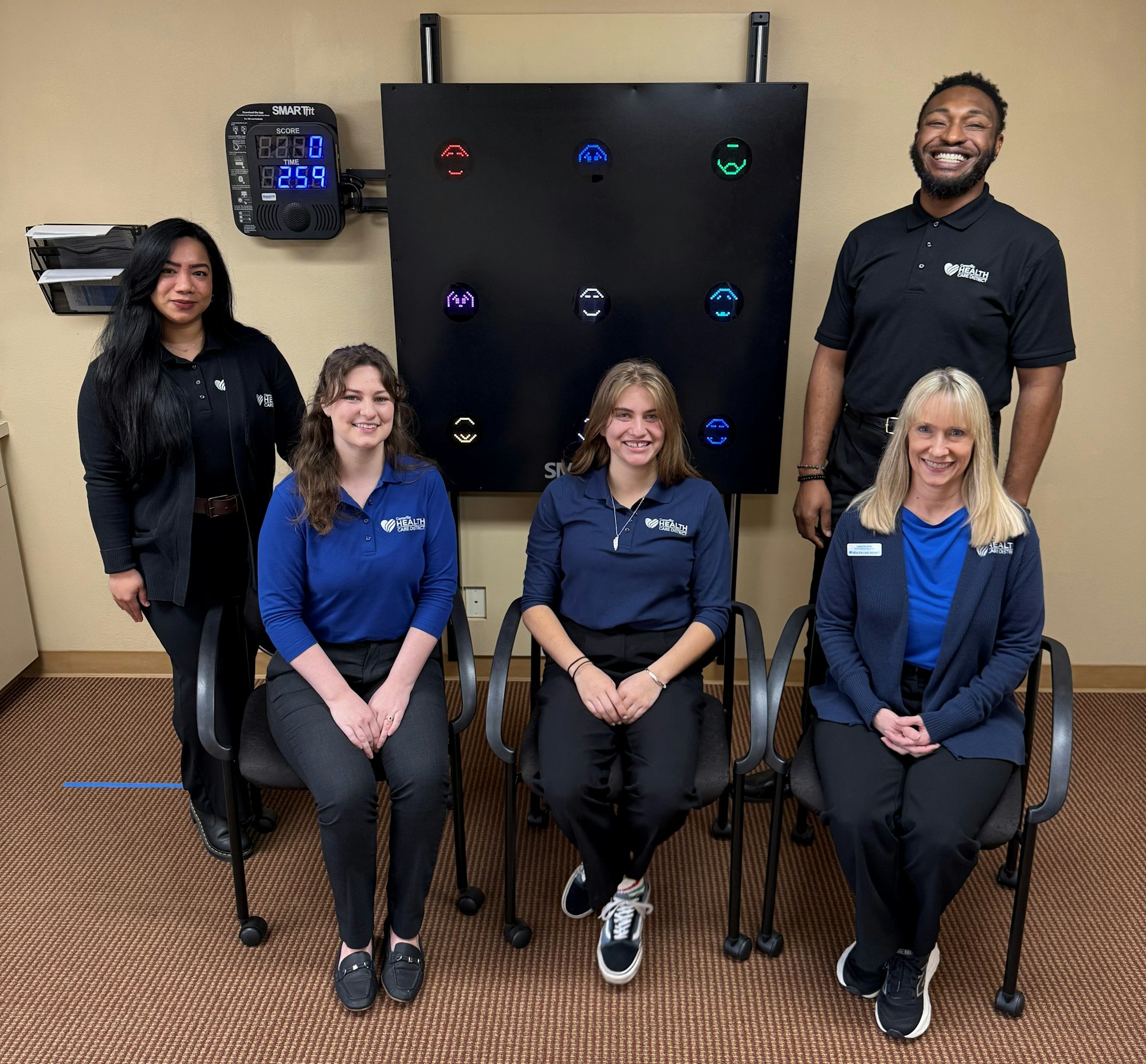 Five people in polo shirts pose together in a clinic or office, with a colorful display board behind them.