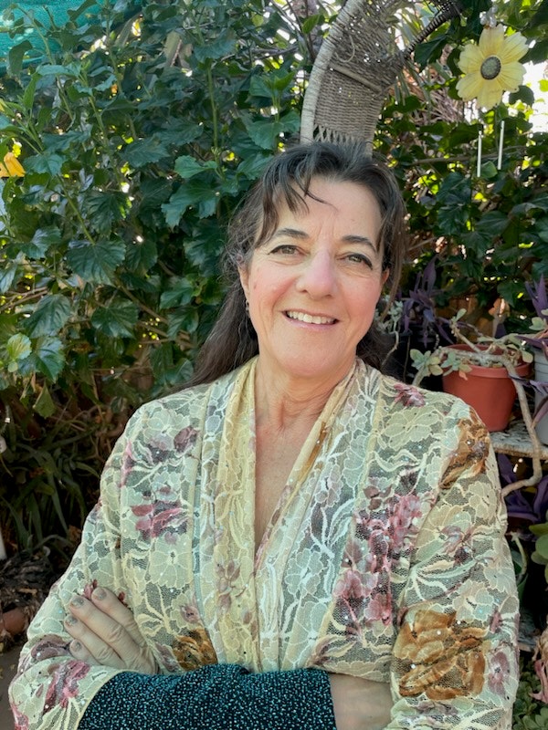 A person smiling, standing in front of leafy plants, wearing a floral-patterned top, and there’s a hanging basket and potted plants nearby.