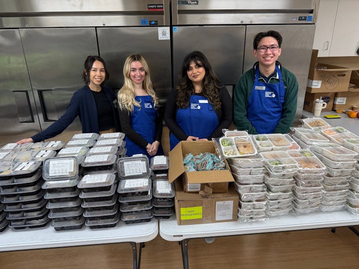 Four people in aprons stand behind a table with stacks of packaged meals.