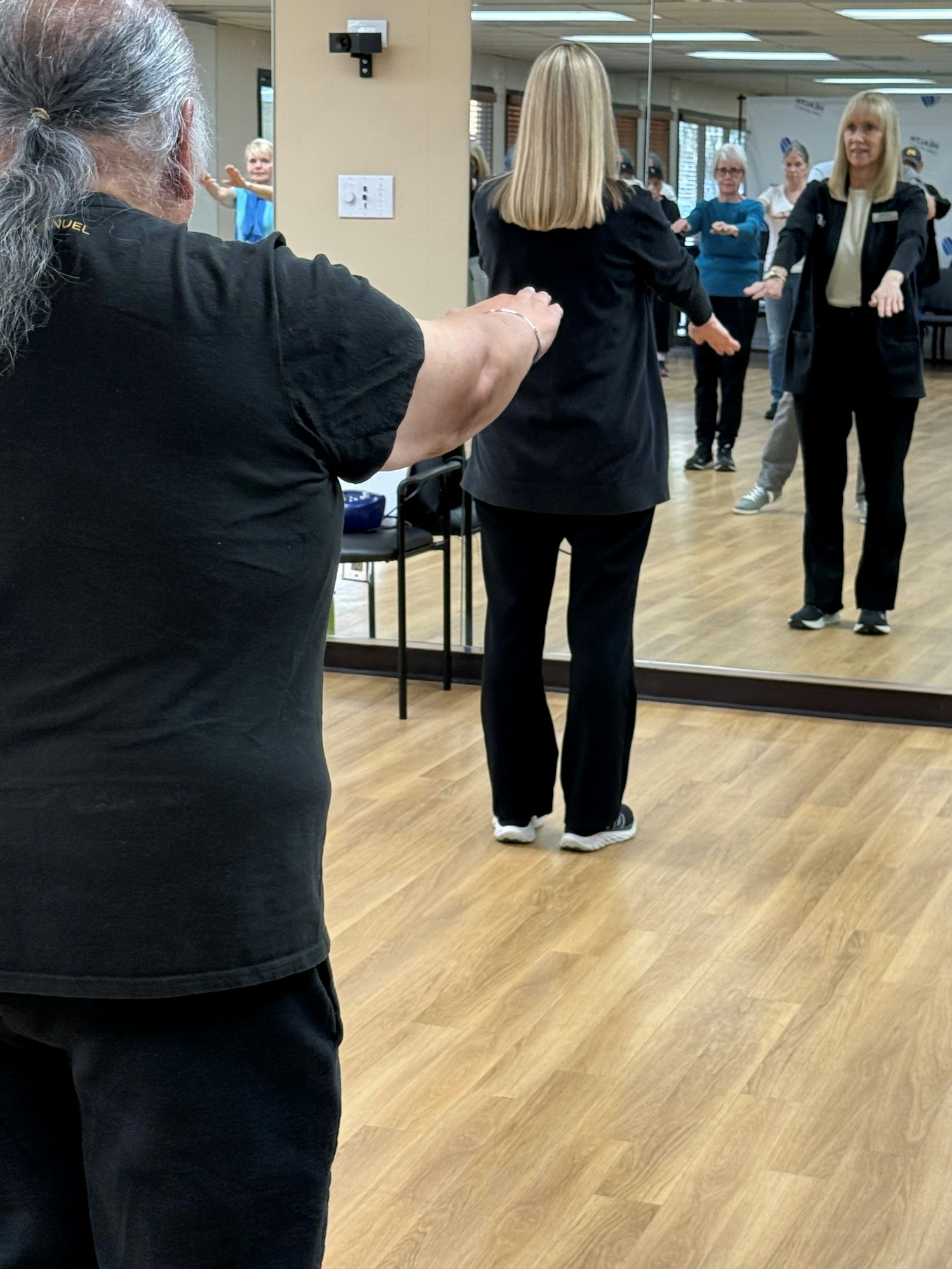 A group session in a studio, with people participating and practicing movements, reflected in a mirror.