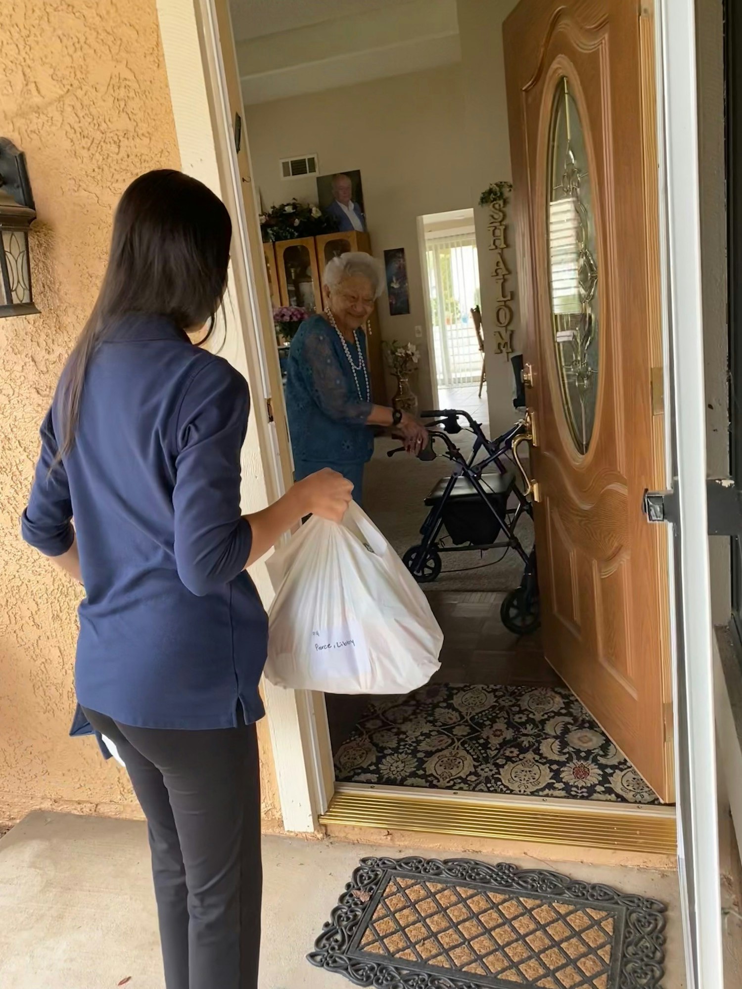 A delivery person handing a bag to an elderly woman using a walker near an open front door.