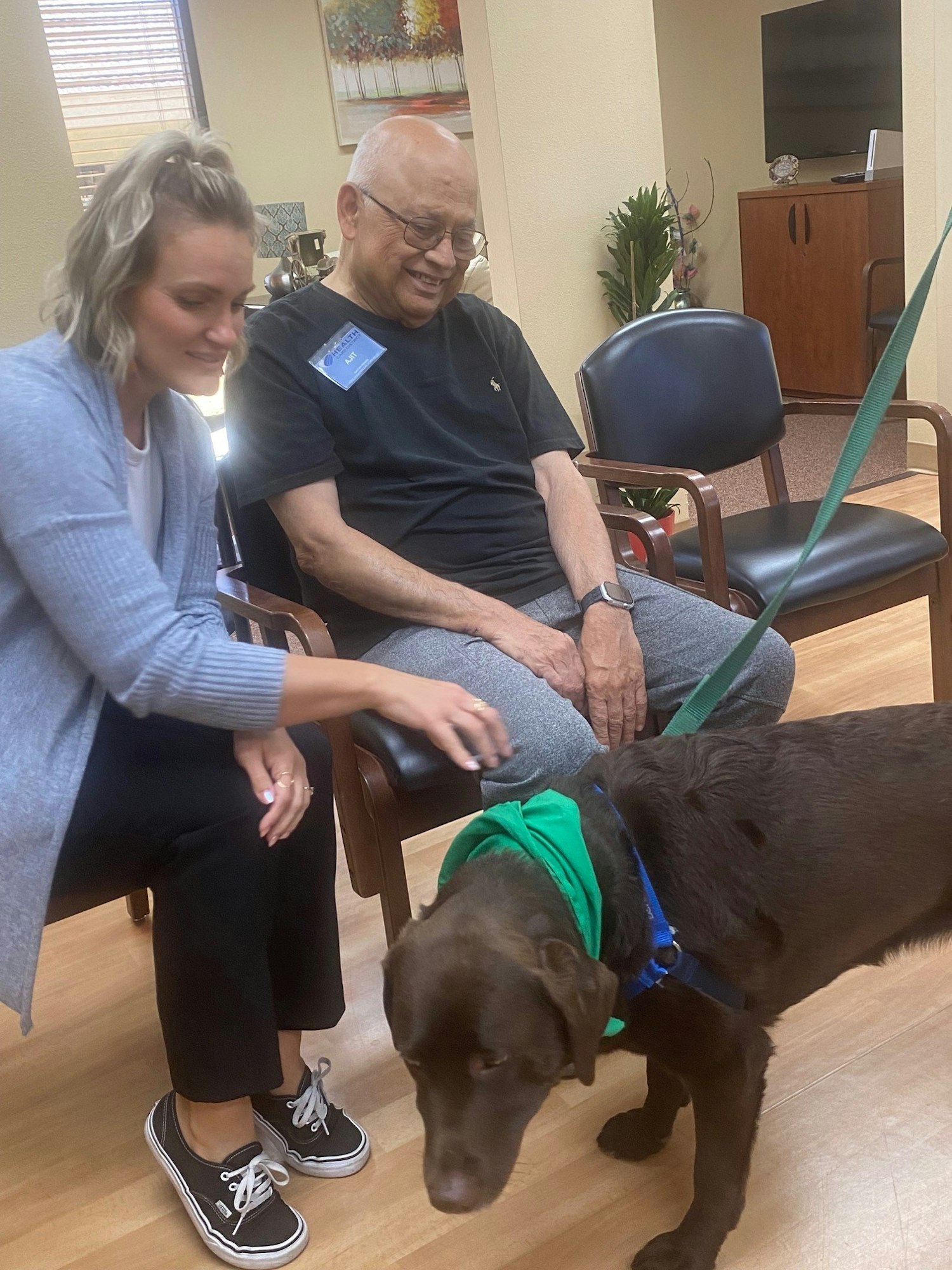 A smiling elderly man sitting, a woman, and a chocolate Labrador in a green vest possibly in a therapy or care setting.