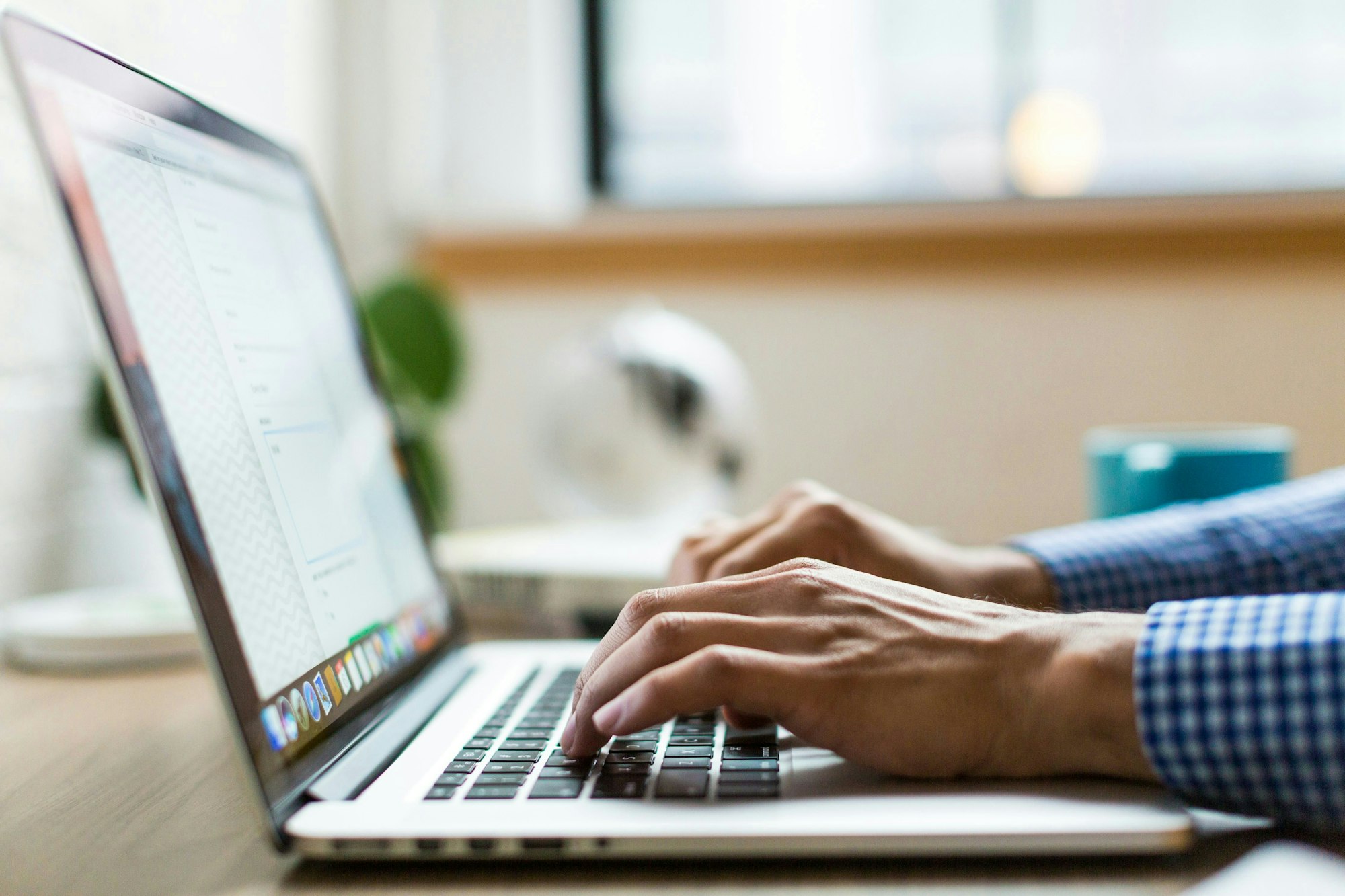 A person typing on a laptop, with a cup and a plant in a bright workspace.