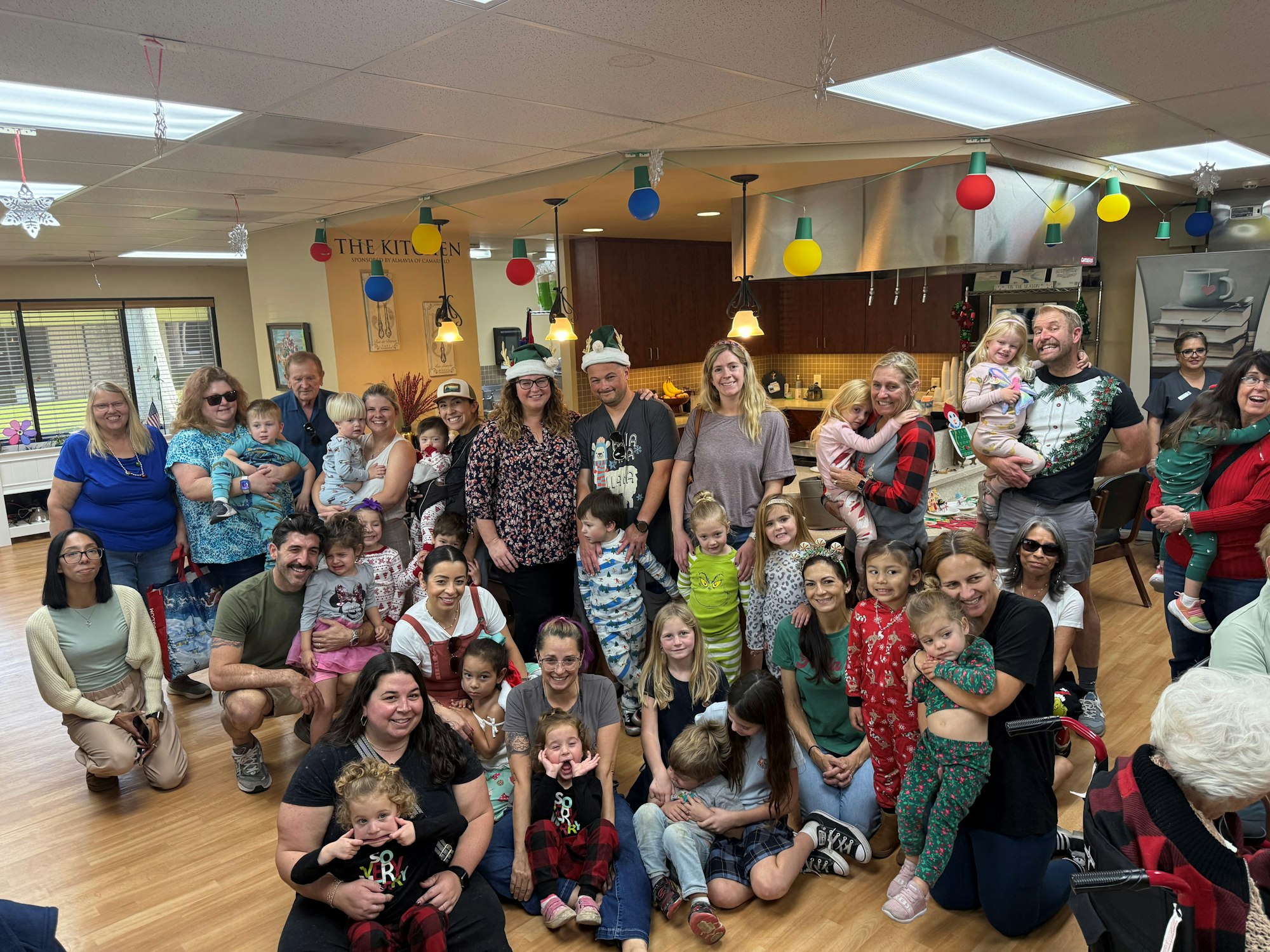 A group of people, including adults and children, gathered indoors, smiling for a photo. Festive decorations are visible in the background.