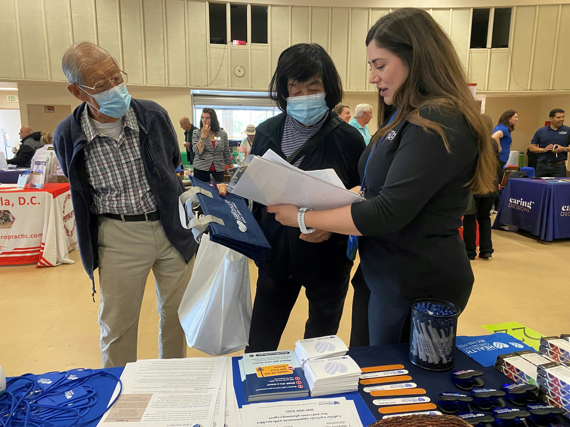 Three people at an indoor event with informational booths, brochures, and freebies on a table.