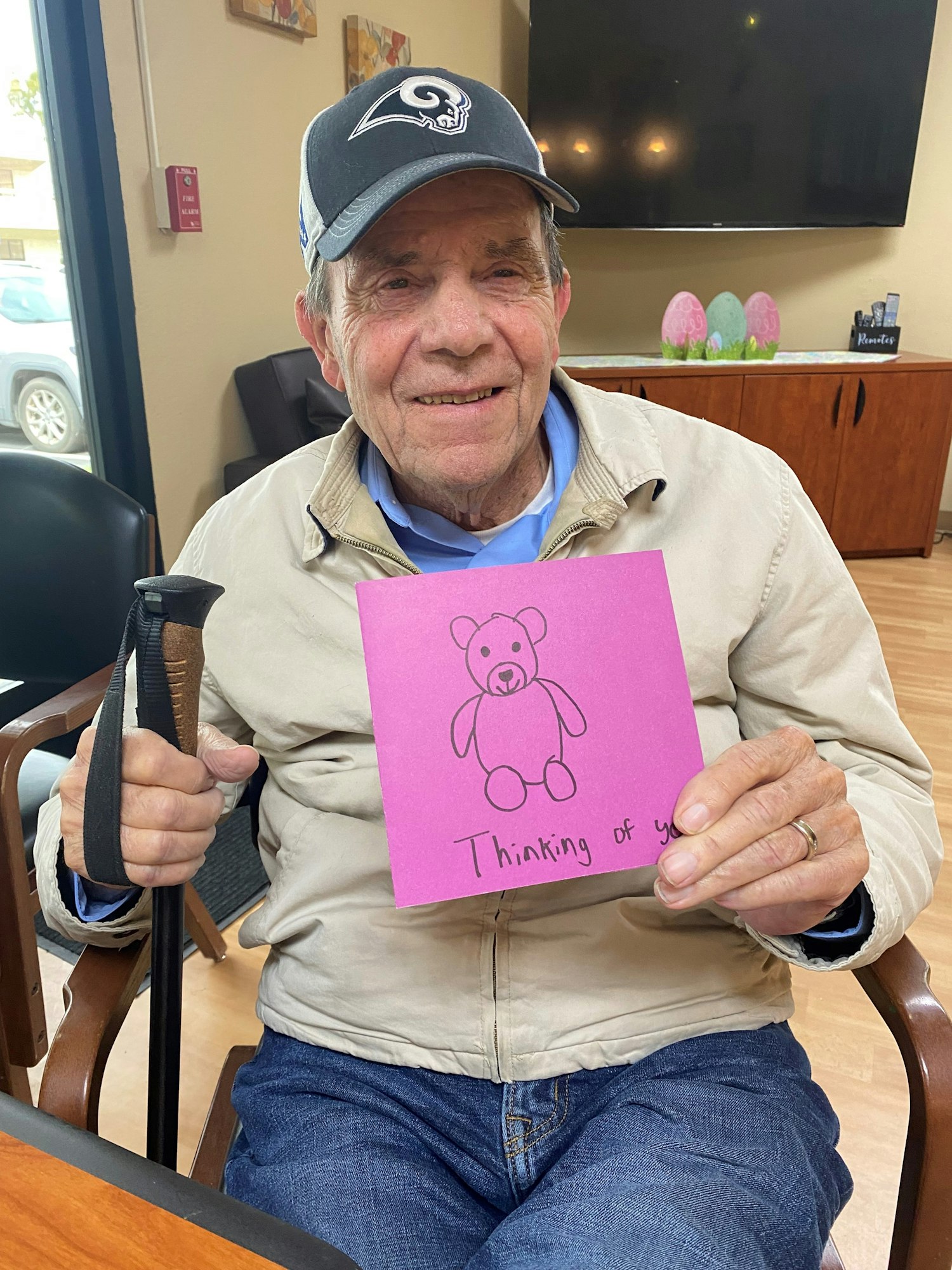 An elderly man in a cap holds a pink card with a teddy bear drawing and the words "Thinking of you."